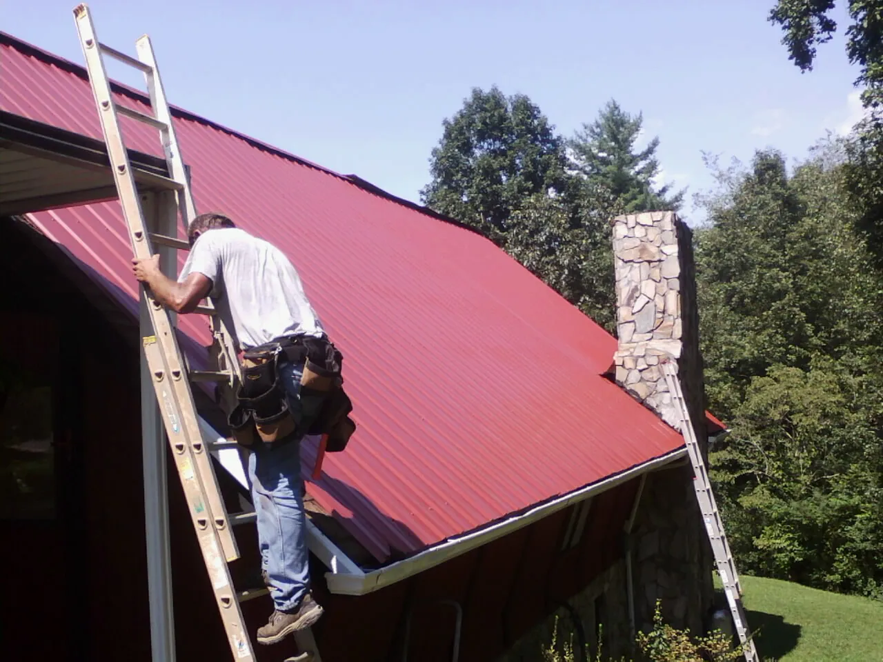 Roofer on a ladder beside a red metal roof and stone chimney — Soffit & Fascia Repair by Roofing Brothers in North Branch